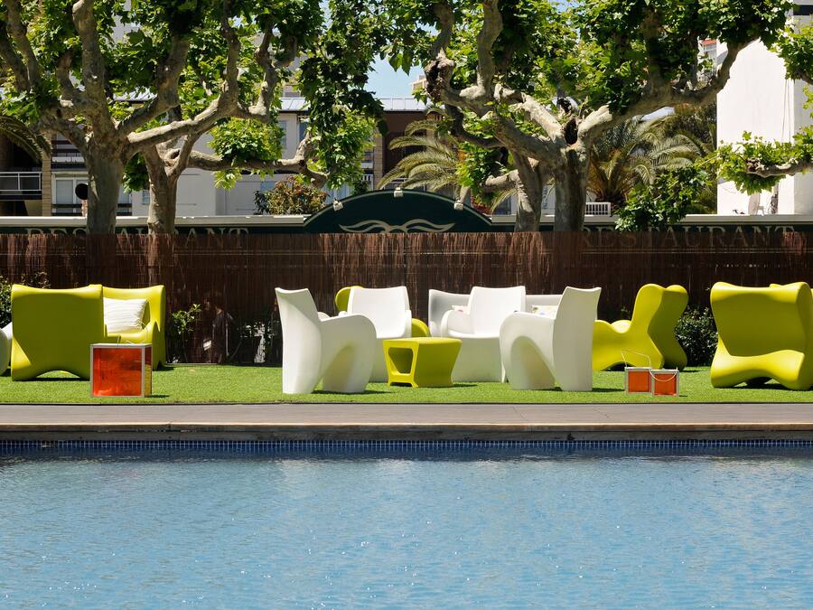 Zona junto a la piscina con modernas sillas reclinables amarillas y blancas, una piscina cristalina y un fondo de árboles y edificios.