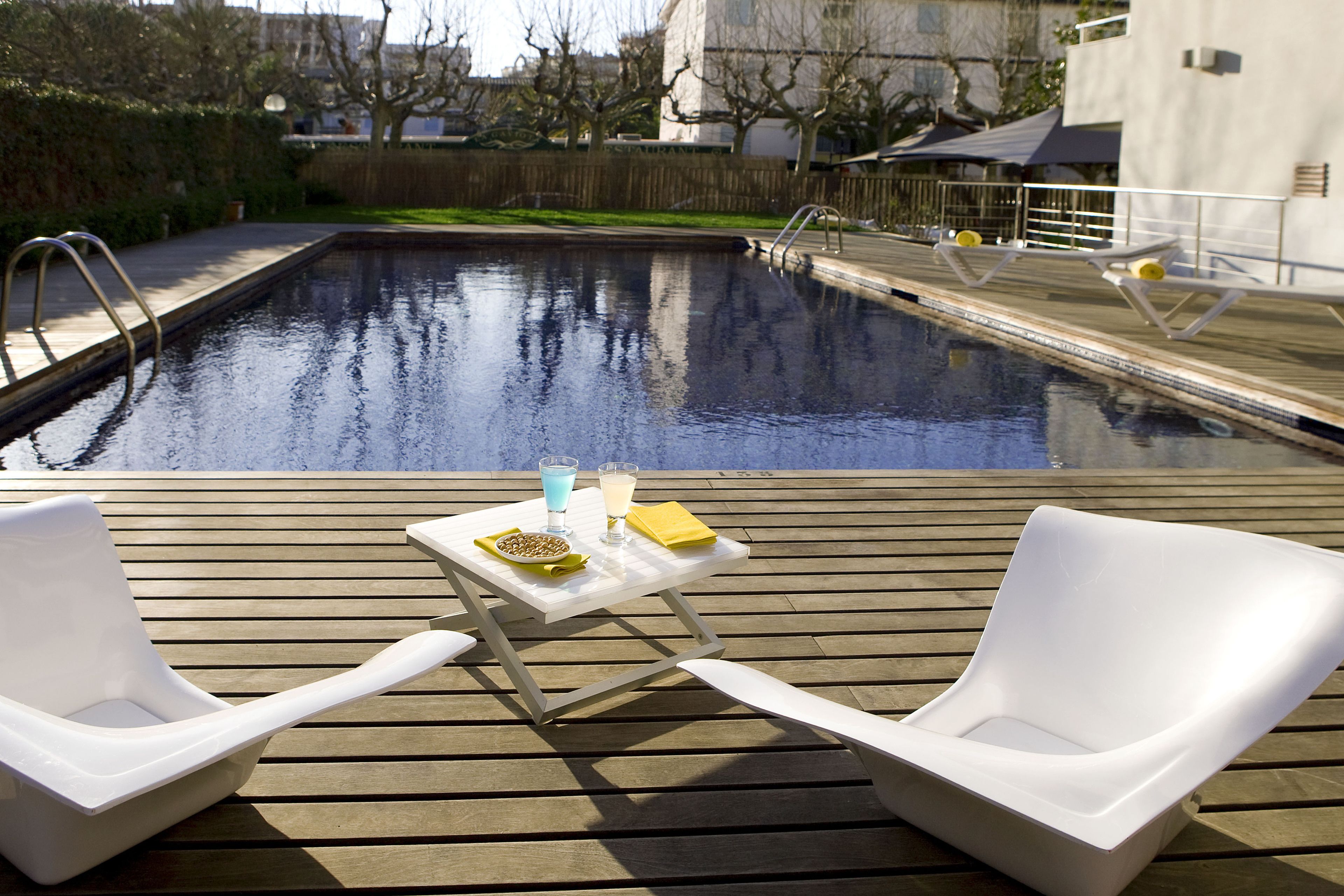 Una piscina con cubierta de madera, dos sillas de playa blancas y una mesa con un vaso, fruta y una servilleta.