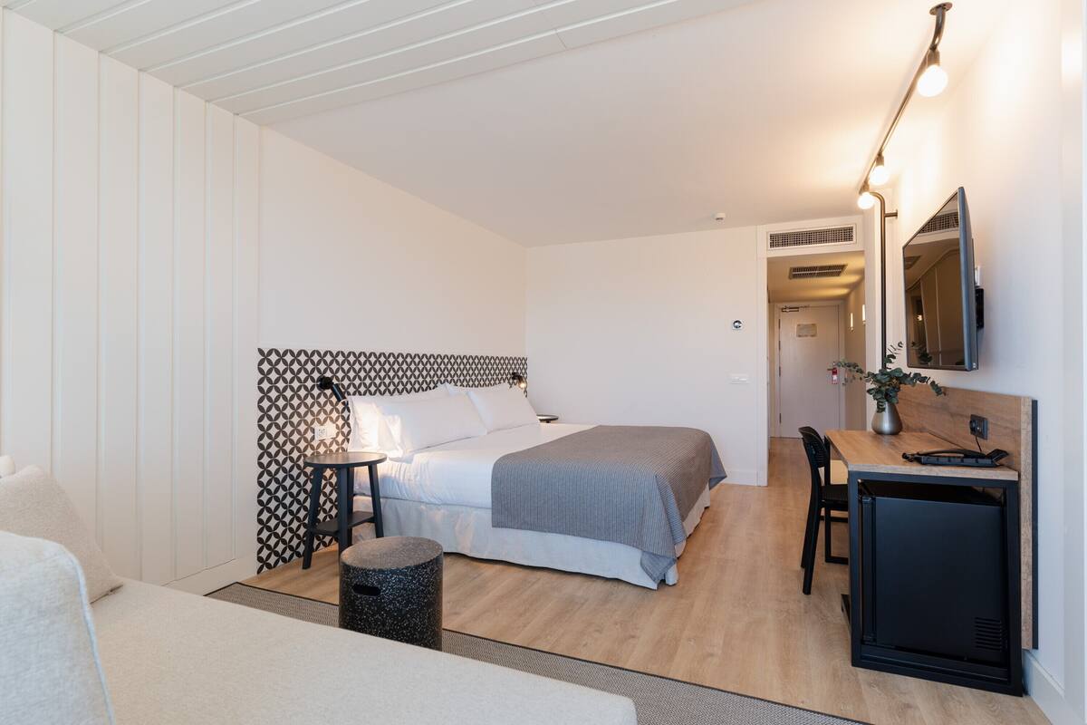 Een moderne hotelkamer met een bed, nachtkastje, lamp, bureau, stoel en een klein zwart kastje.