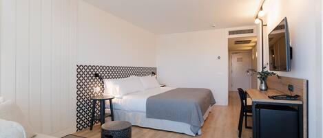 A modern hotel room with a bed, bedside table, lamp, desk, chair, and a small black cabinet.