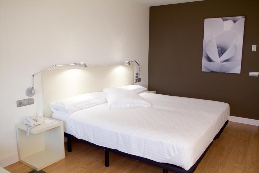 Een bed met wit beddengoed, een nachtkastje met een lamp en een kunstwerk aan de muur.