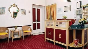 Une chambre d’hôtel avec un tapis rouge, deux fauteuils, une petite table, une bibliothèque et une porte ornée de rideaux rouges.