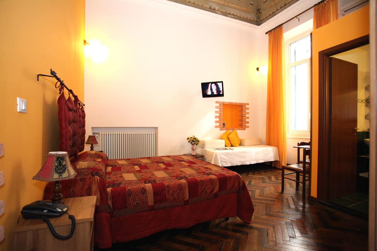 Una habitación de hotel con una cama grande, una mesita de noche con una lámpara, una pequeña mesa con un florero y un televisor en la pared.