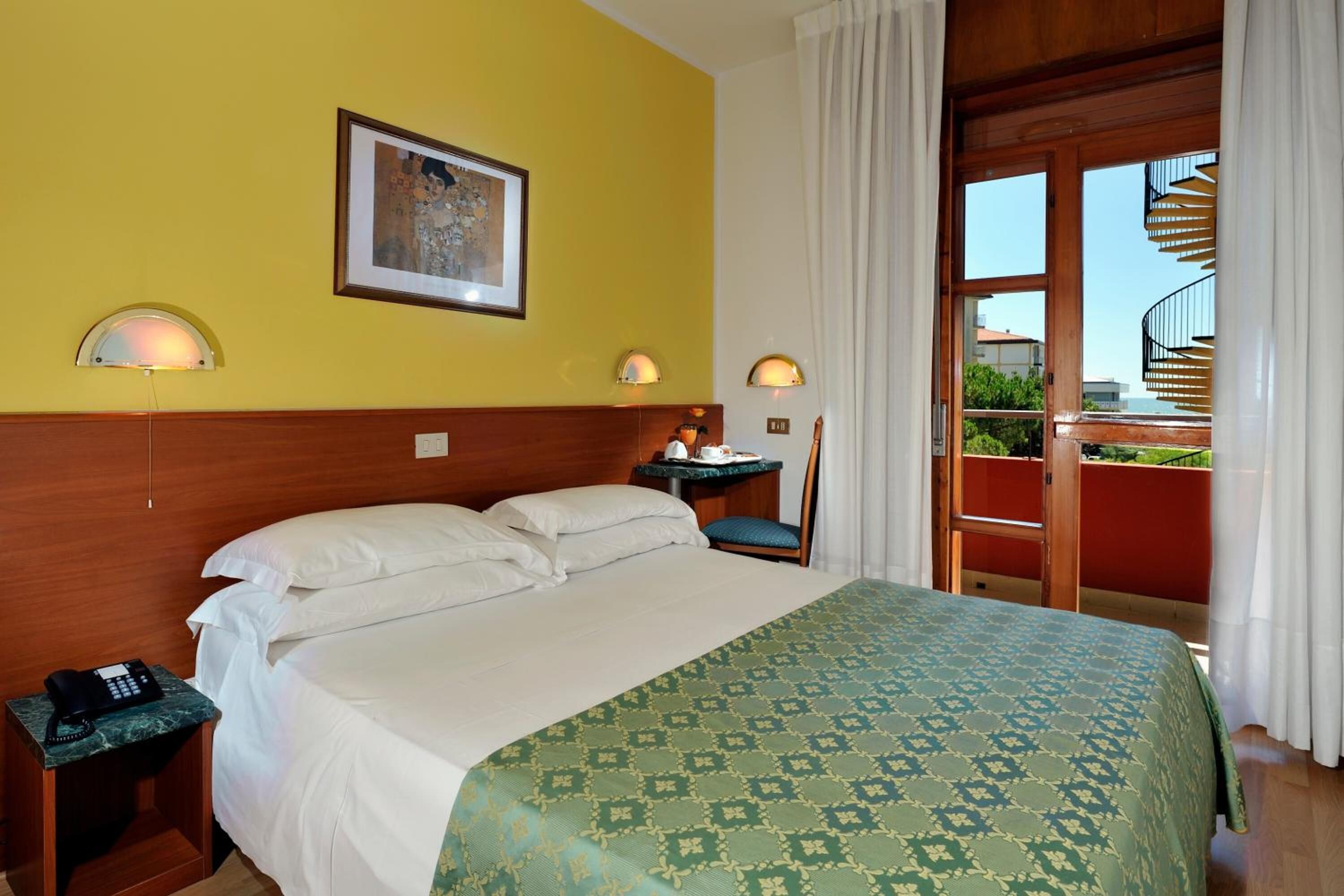 A hotel room with a bed, a nightstand, a chair, a balcony with a railing, and a painting on the wall.