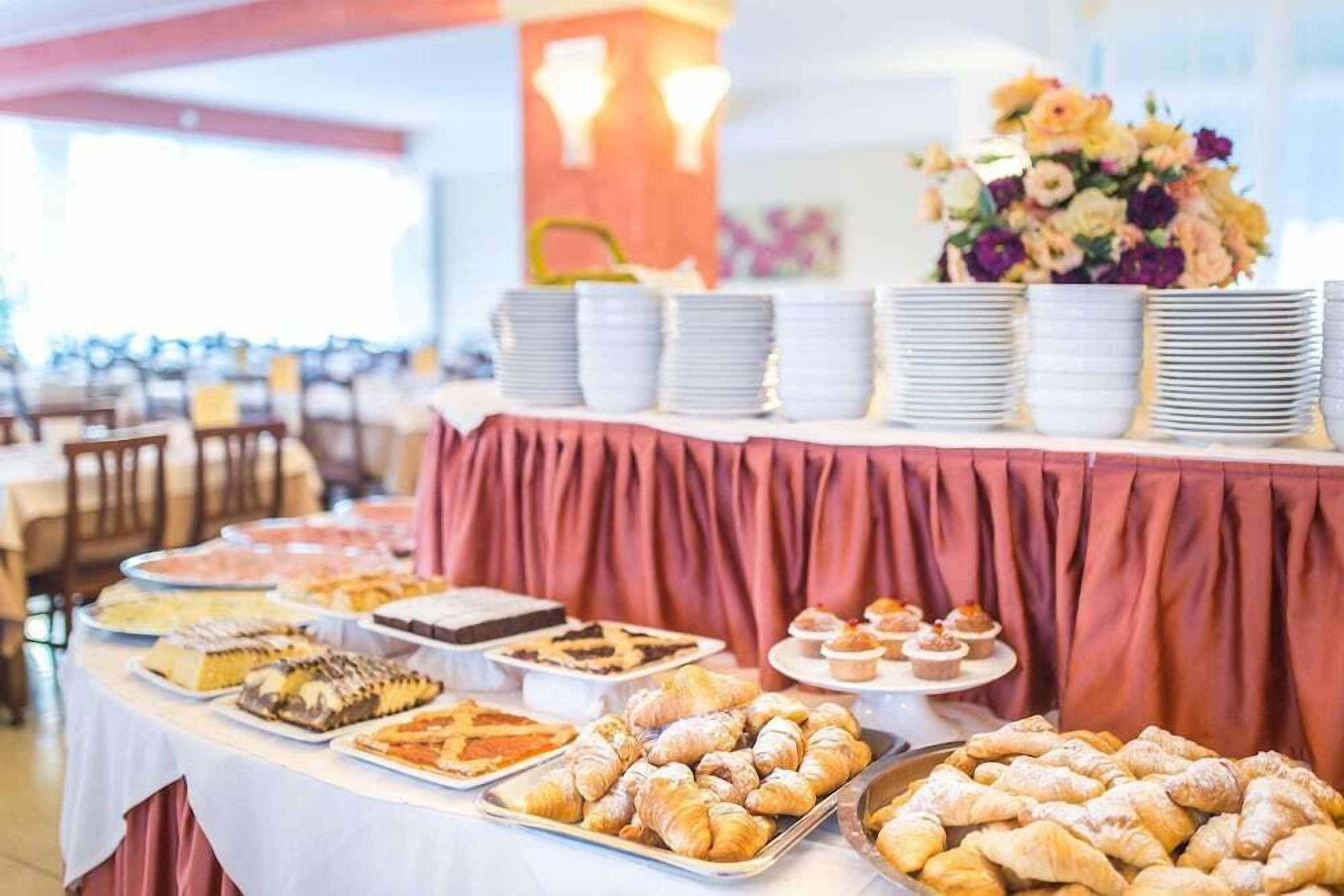 Ein reich gedeckter Buffet mit verschiedenen Gebäcken und Desserts, dazu gestapelten Tellern und einer Blumenkomposition im Hintergrund.