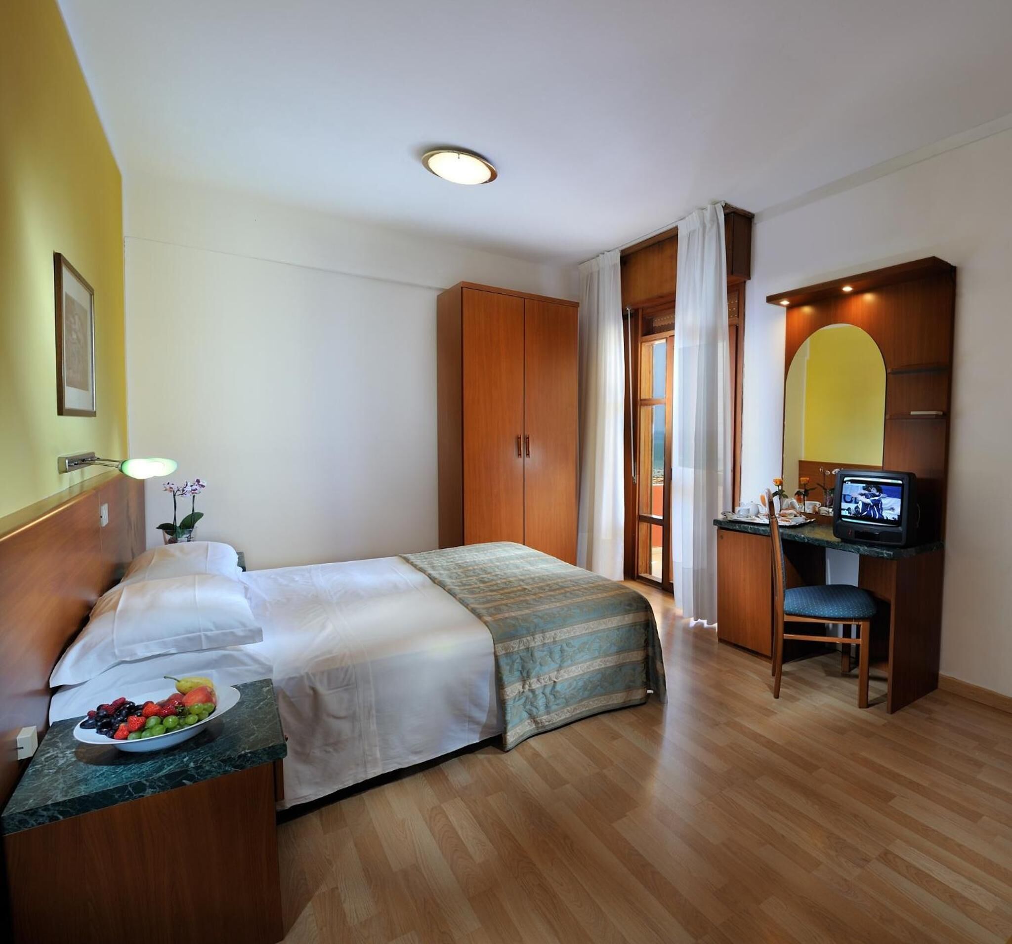 Ein Hotelzimmer mit einem Bett, einem Schreibtisch mit Computer, einem Stuhl und einem kleinen Tisch mit Obst.