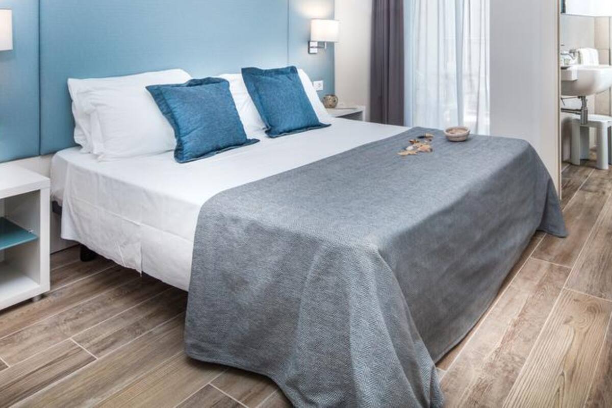 Une chambre d’hôtel moderne avec un grand lit, deux oreillers, une couverture grise, une table de chevet, une commode, un miroir et une salle de bain visible à travers une porte ouverte.