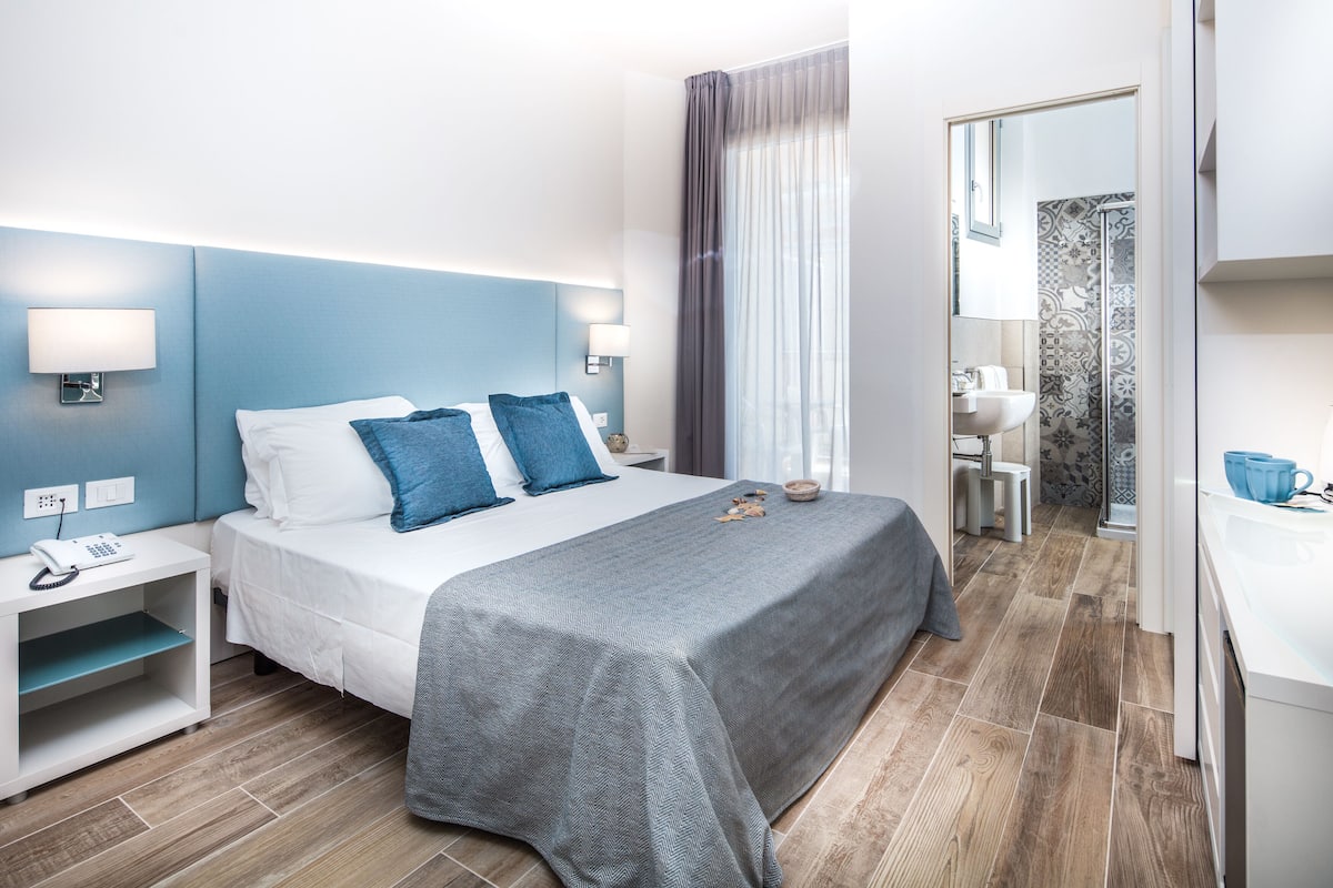 Une chambre d’hôtel moderne équipée d’un lit, de tables de chevet, d’une petite table, d’une salle de bain avec un lavabo et un miroir, et d’un espace douche.