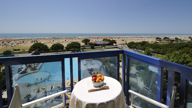 A balcony with a table holding a bowl of fruit, overlooking a beach with umbrellas and a swimming pool. - Hotel Bellevue (San Michele al Tagliamento)