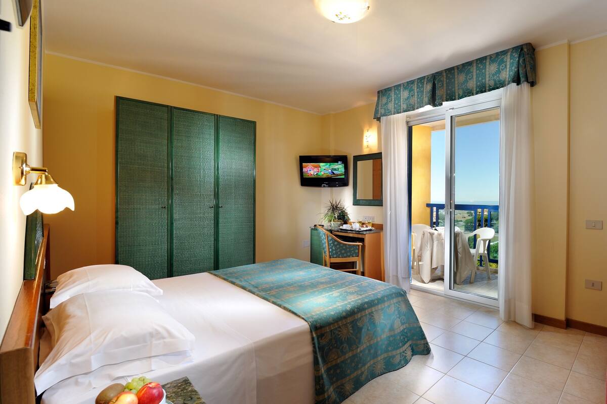 Een hotelkamer met een bed, een bureau met stoel, een televisie en een balkon met uitzicht.