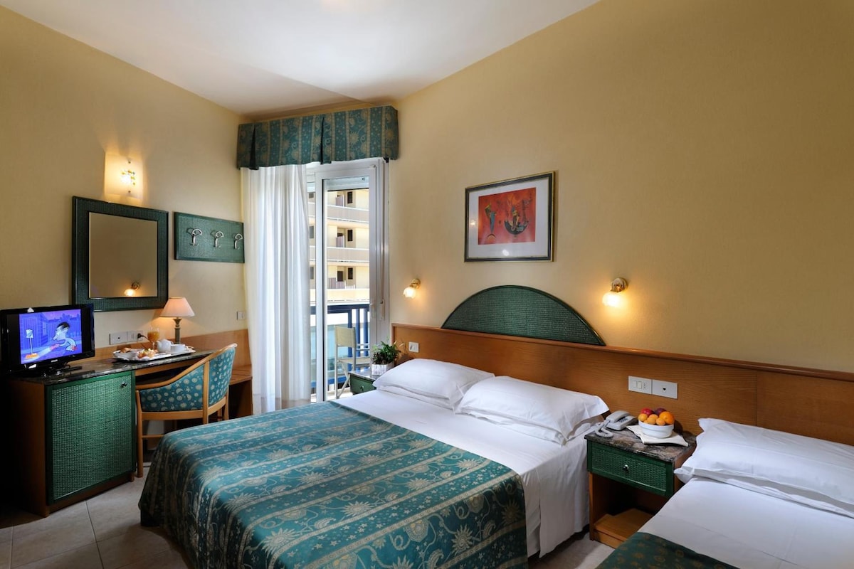 Ein Hotelzimmer mit Bett, Schreibtisch mit Fernseher, Stuhl, Nachttisch und Fenster mit Blick auf ein Gebäude.