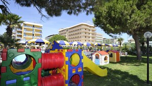 A playground with colorful play structures, a small house, and a building with balconies in the background. - Hotel Bellevue (San Michele al Tagliamento)