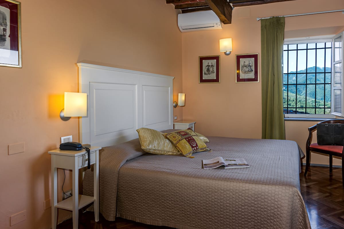Un lit bien fait, recouvert d’une courtepointe, une table de chevet avec une lampe et une fenêtre donnant sur l’extérieur.