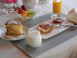 Un desayuno con una porción de pastel, un bol de fruta, un vaso de zumo de naranja y un frasco pequeño de nata.