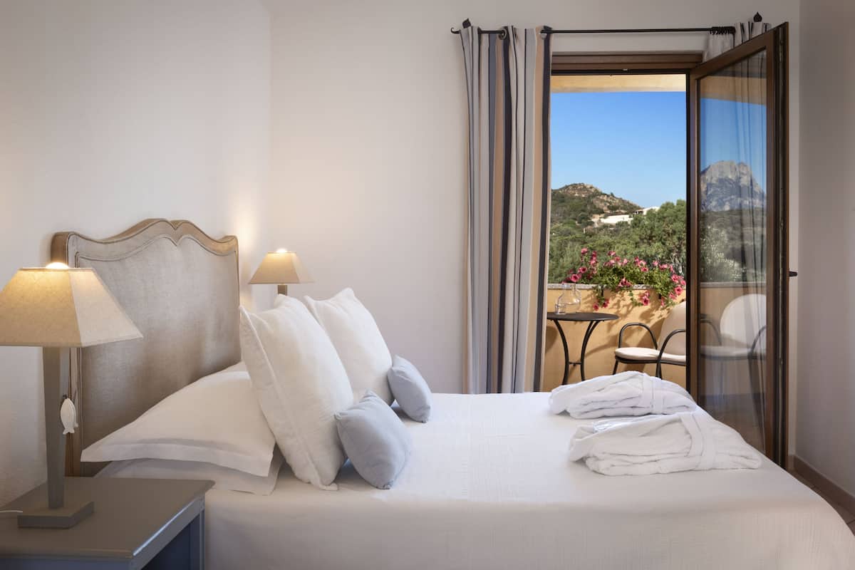 Ein Hotelzimmer mit Bett, zwei Nachttischlampen, Blick auf einen Balkon mit Tisch und Stühlen sowie einem Berg im Hintergrund.