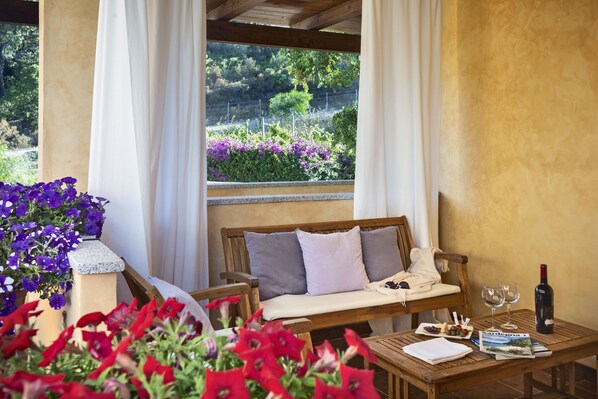 Un banco de madera con cojines, una mesa con una botella de vino y copas, y una ventana con vista a un jardín.
