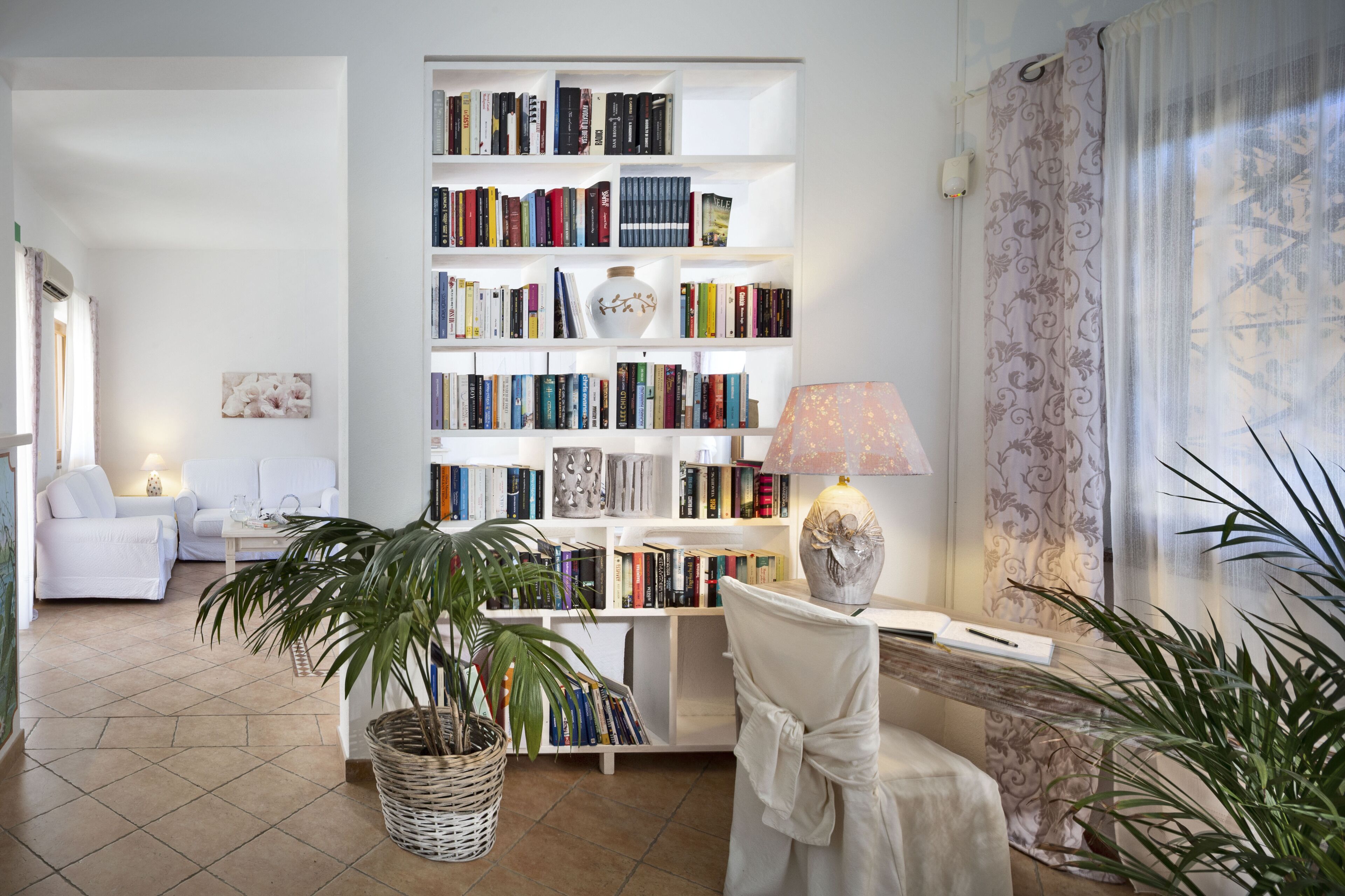 Een kamer met een grote boekenkast, een witte stoel, een lamp en een potplant.