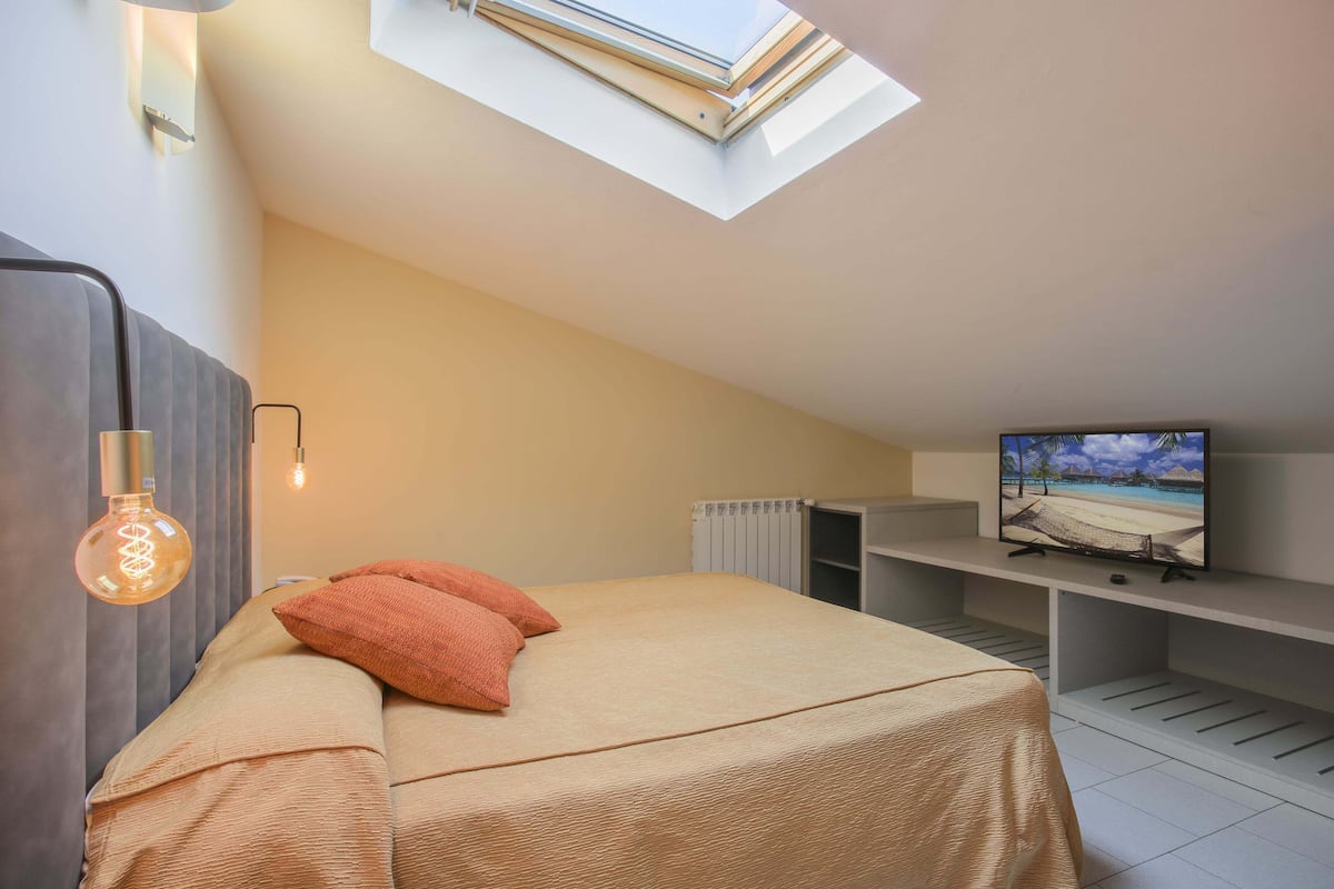 Ein Schlafzimmer mit Bett, Fernseher auf einem Schrank, Oberlicht und Heizkörper.