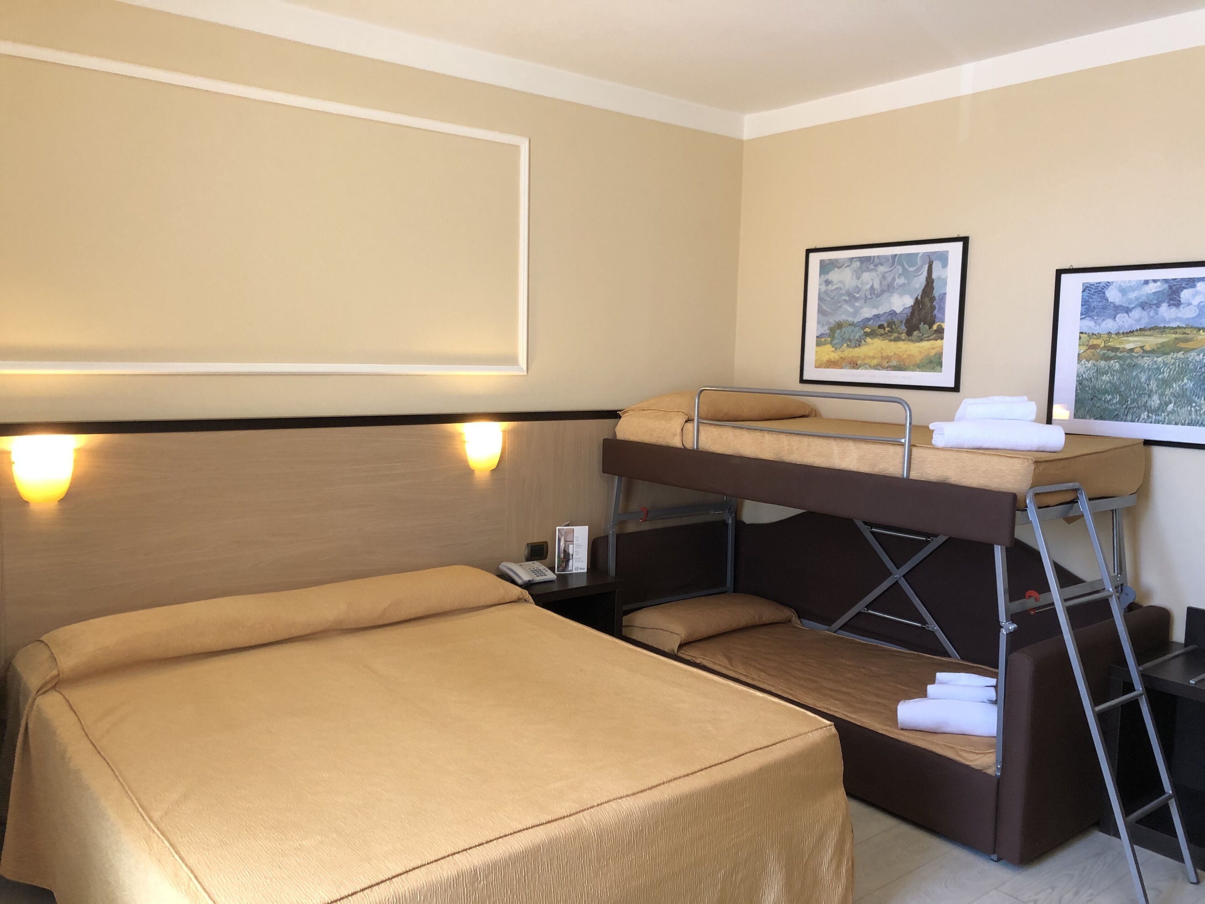 Quarto de hotel com uma cama de solteiro, uma escrivaninha e duas imagens emolduradas na parede.