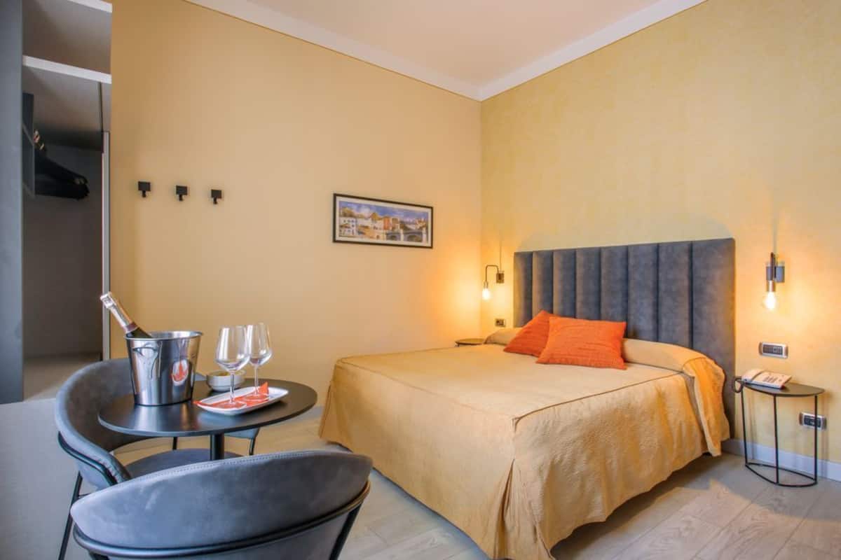 Ein Hotelzimmer mit Bett, einem kleinen Tisch mit Flasche und Gläsern, einem Sessel und einem Nachttisch mit Lampe.