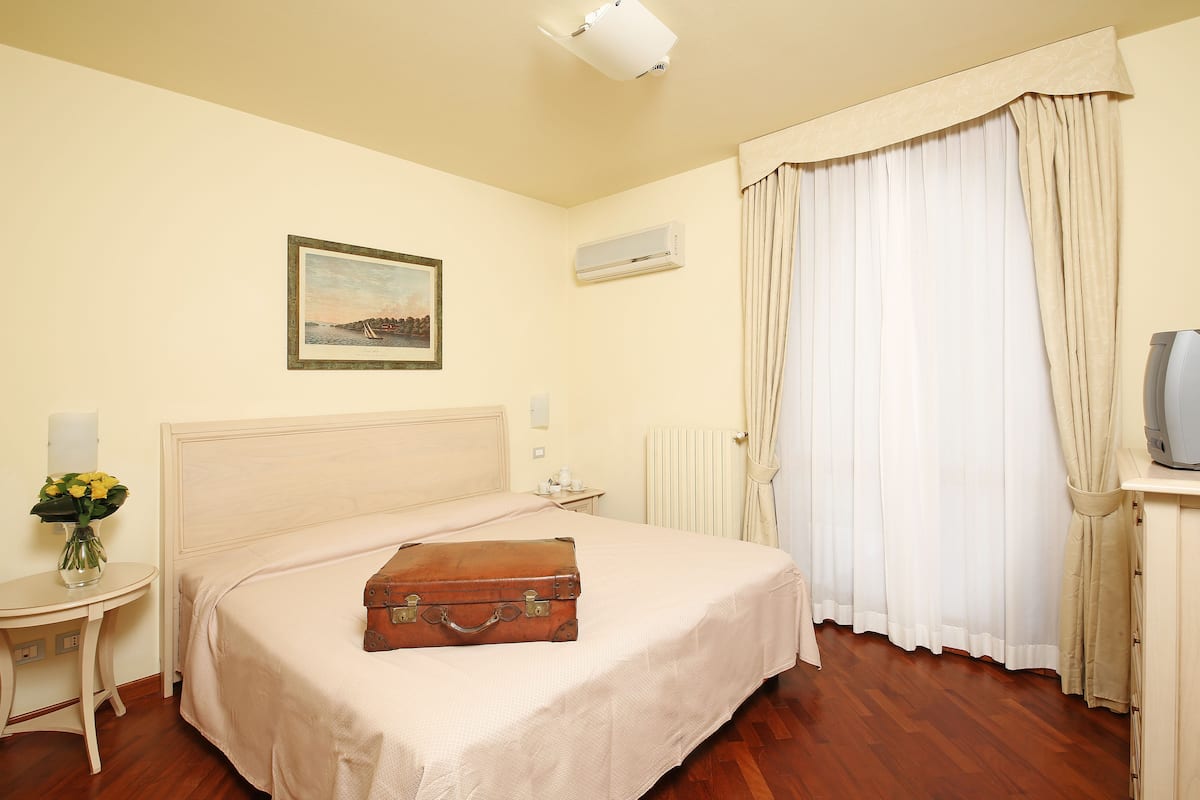 A hotel room with a bed, a bedside table with a vase of flowers, an air conditioning unit, a radiator, and a window with curtains.