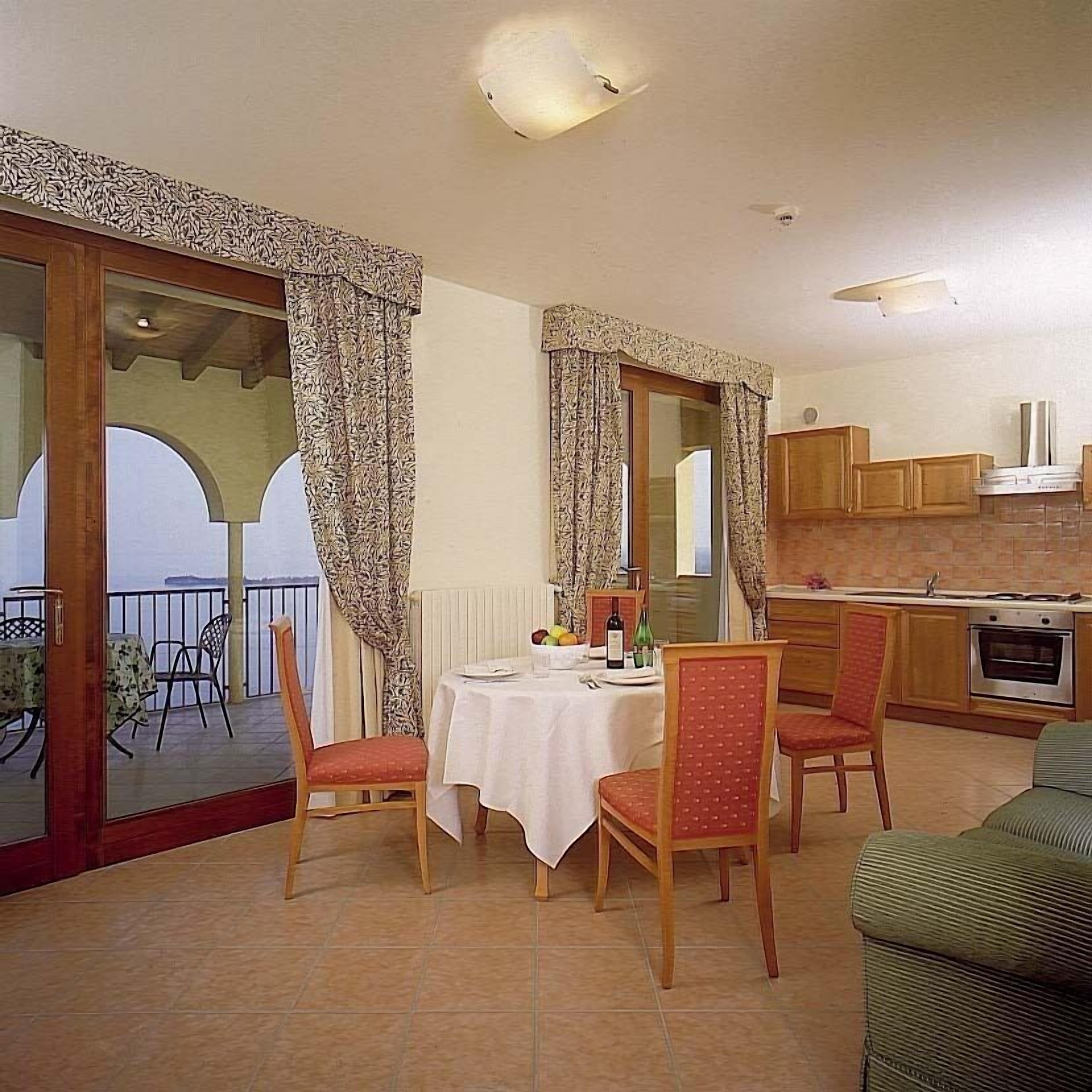 A dining area with a table set for two, chairs, a view of the outside, and a kitchen area with cabinets and appliances.
