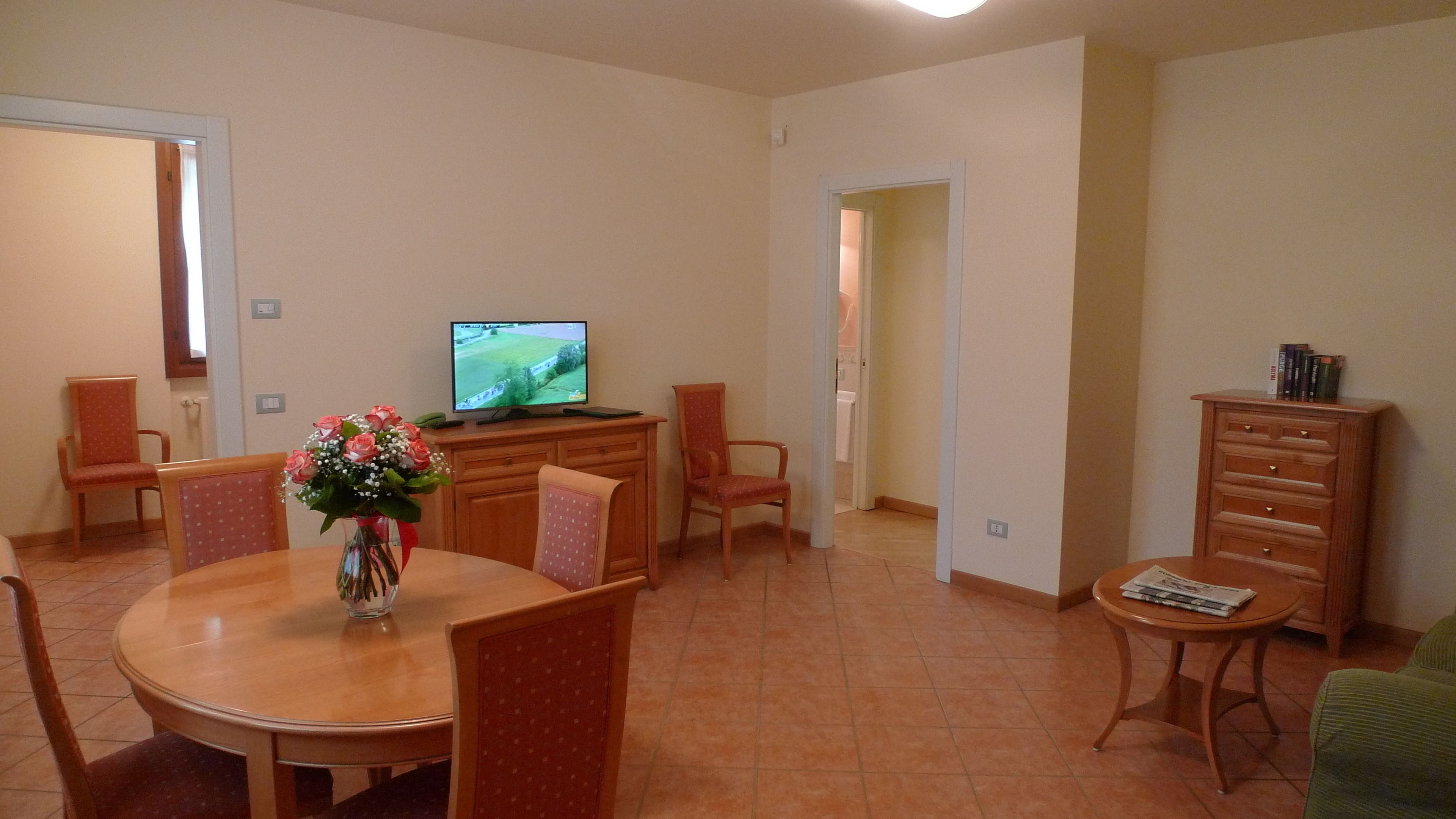 Un comedor con mesa de madera, sillas, un florero, un televisor y un mueble con libros.