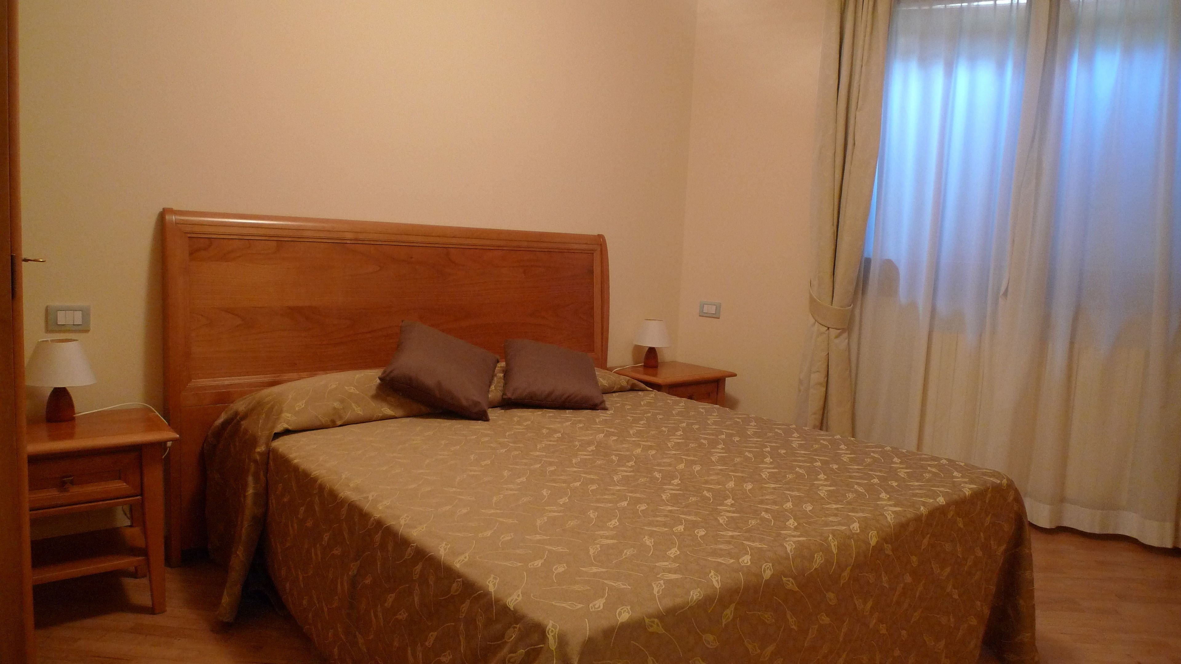 A standard hotel room with a wooden bed, two pillows, a nightstand with a lamp, and a window with curtains.