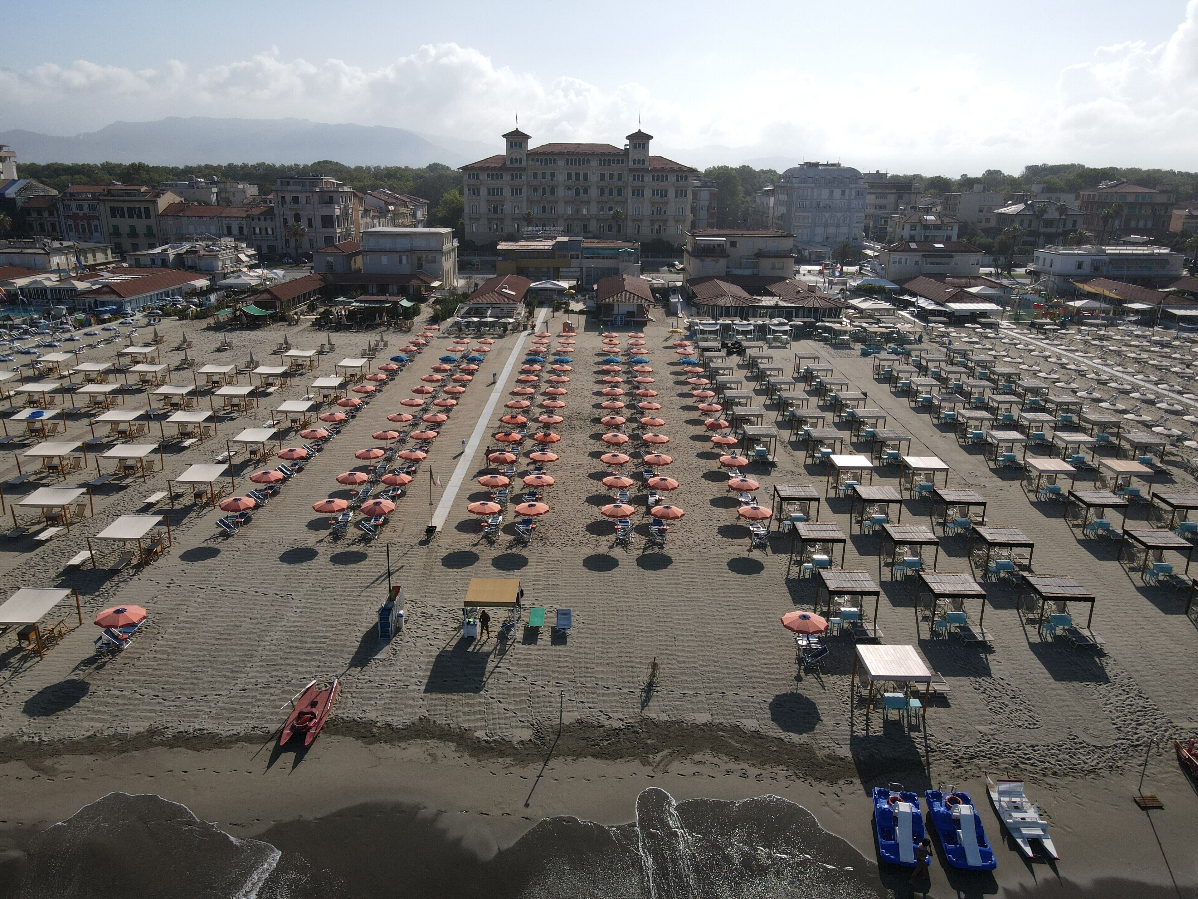 A beach with rows of sunbeds and umbrellas, a few boats, and buildings in the background.