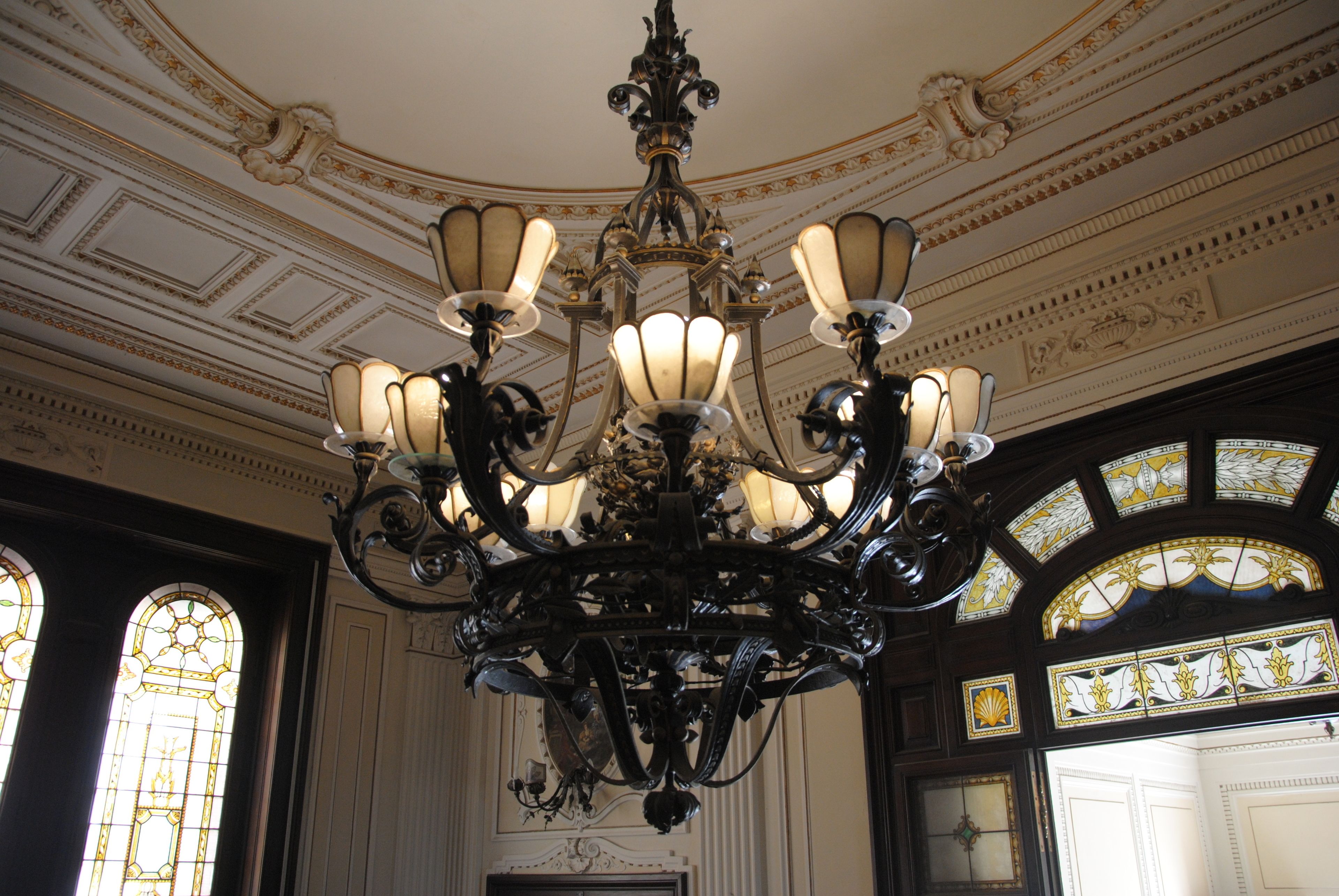 An ornate chandelier with multiple light fixtures, a detailed ceiling with decorative moldings, and large stained glass windows.