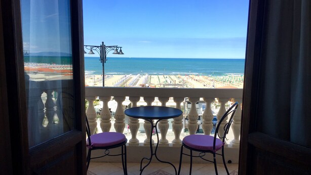 Un balcón con dos sillas y una mesita pequeña, con vistas a una playa con sombrillas y cielo despejado.