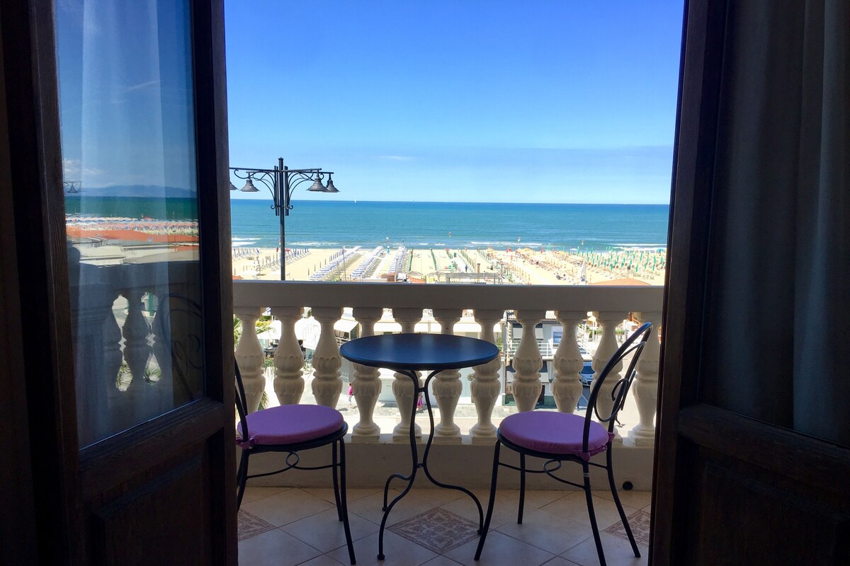 A balcony with two chairs and a small table, overlooking a beach with umbrellas and a clear sky.