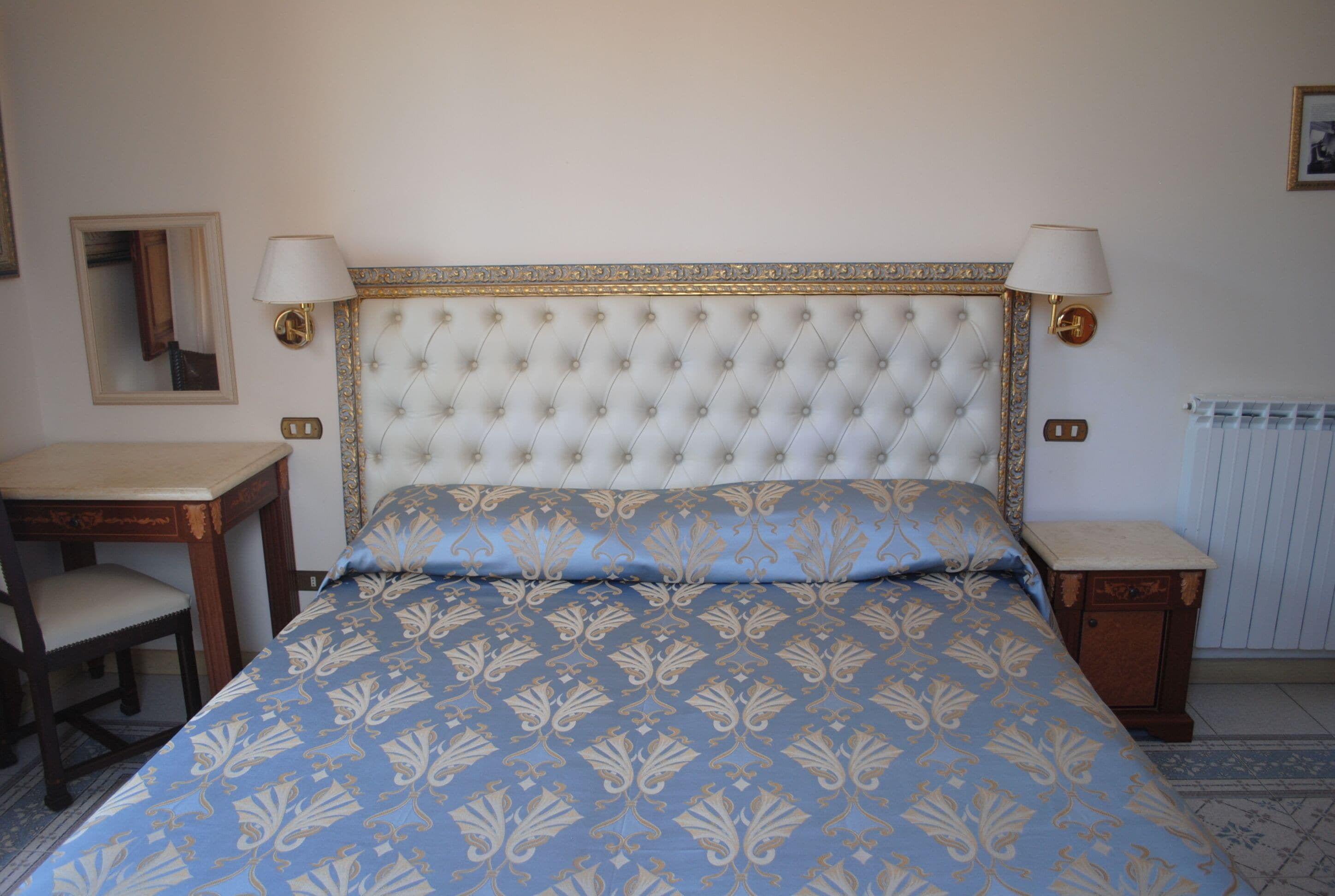 Un dormitorio con cabecera capitonada, dos lámparas de noche, cama con edredón estampado, mesita de noche, una silla y un radiador.
