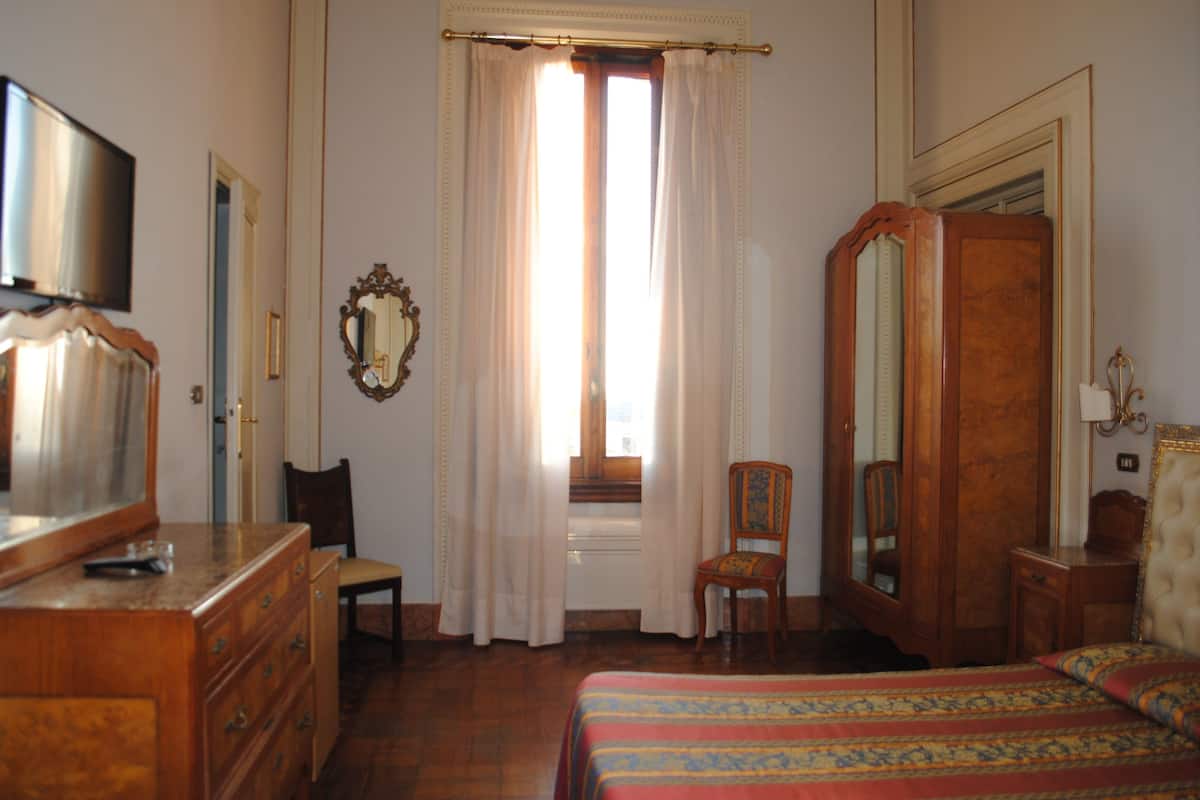 A bedroom with a bed, a wooden dresser, a chair, a wardrobe, a mirror, and a television.