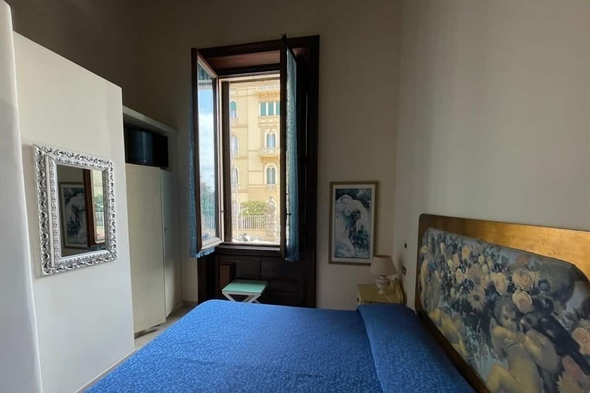 A bedroom with a bed, a window, a small table, and a painting on the wall.