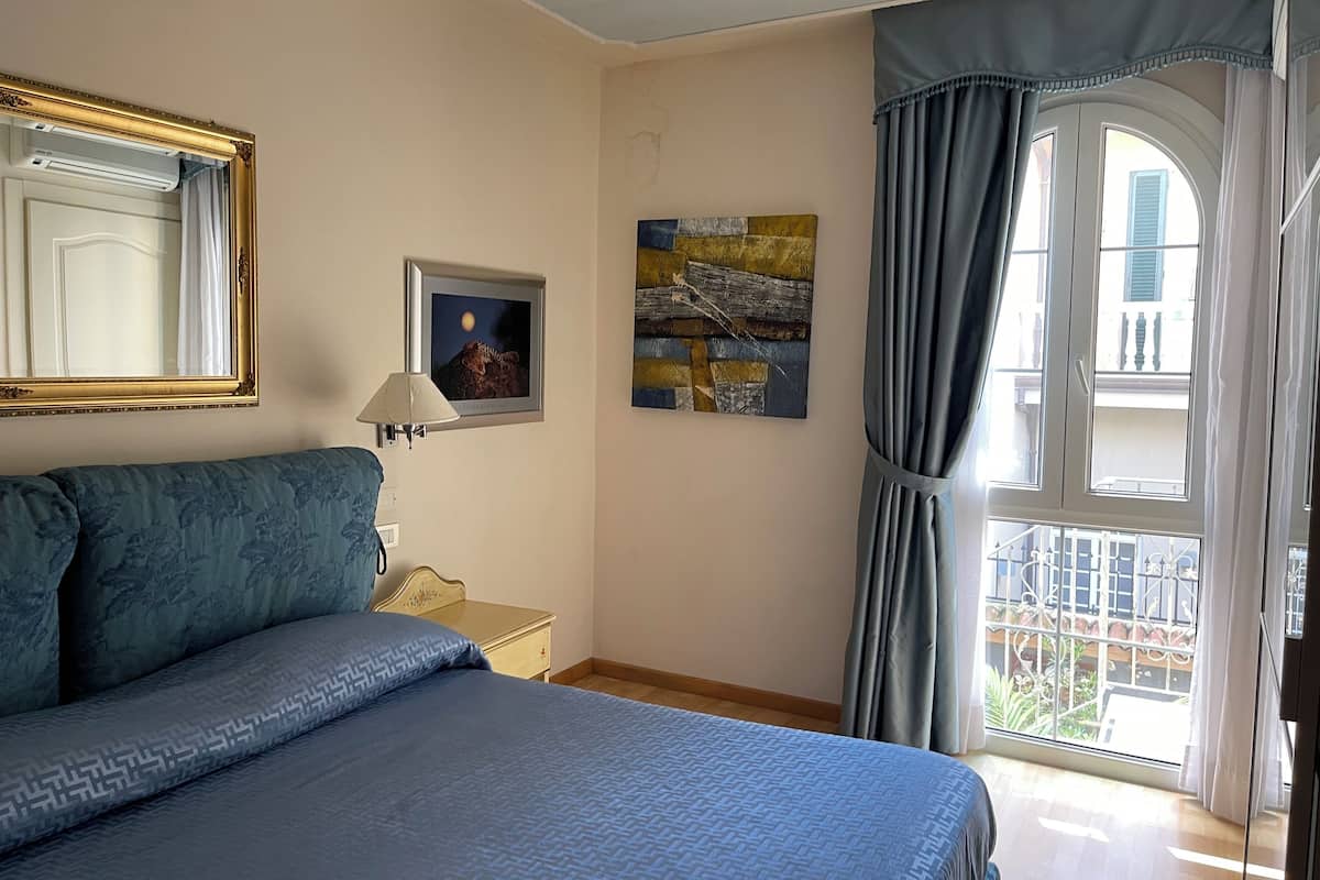 A bedroom with a bed, a nightstand, a lamp, a framed picture, and a window with curtains.
