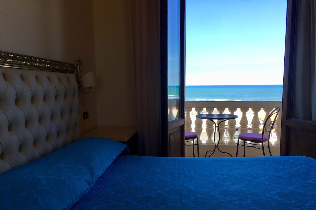 A hotel room with a bed, a view of the beach, and a balcony with two chairs.