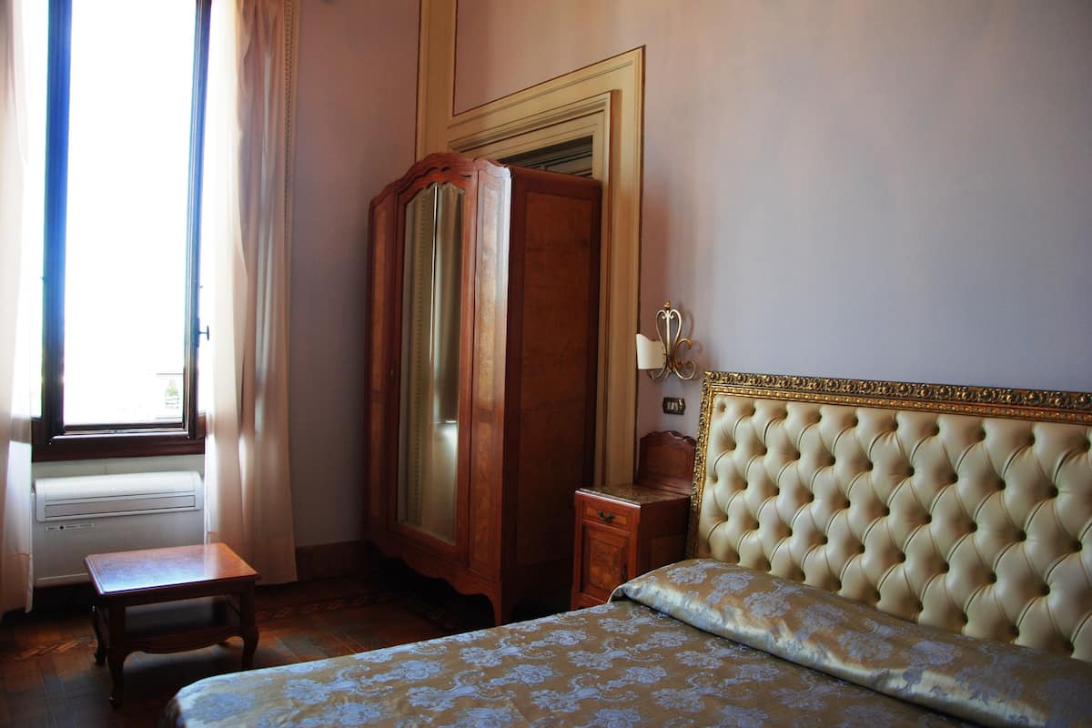A bedroom with a bed, a wooden wardrobe, a small table, and a window with curtains.