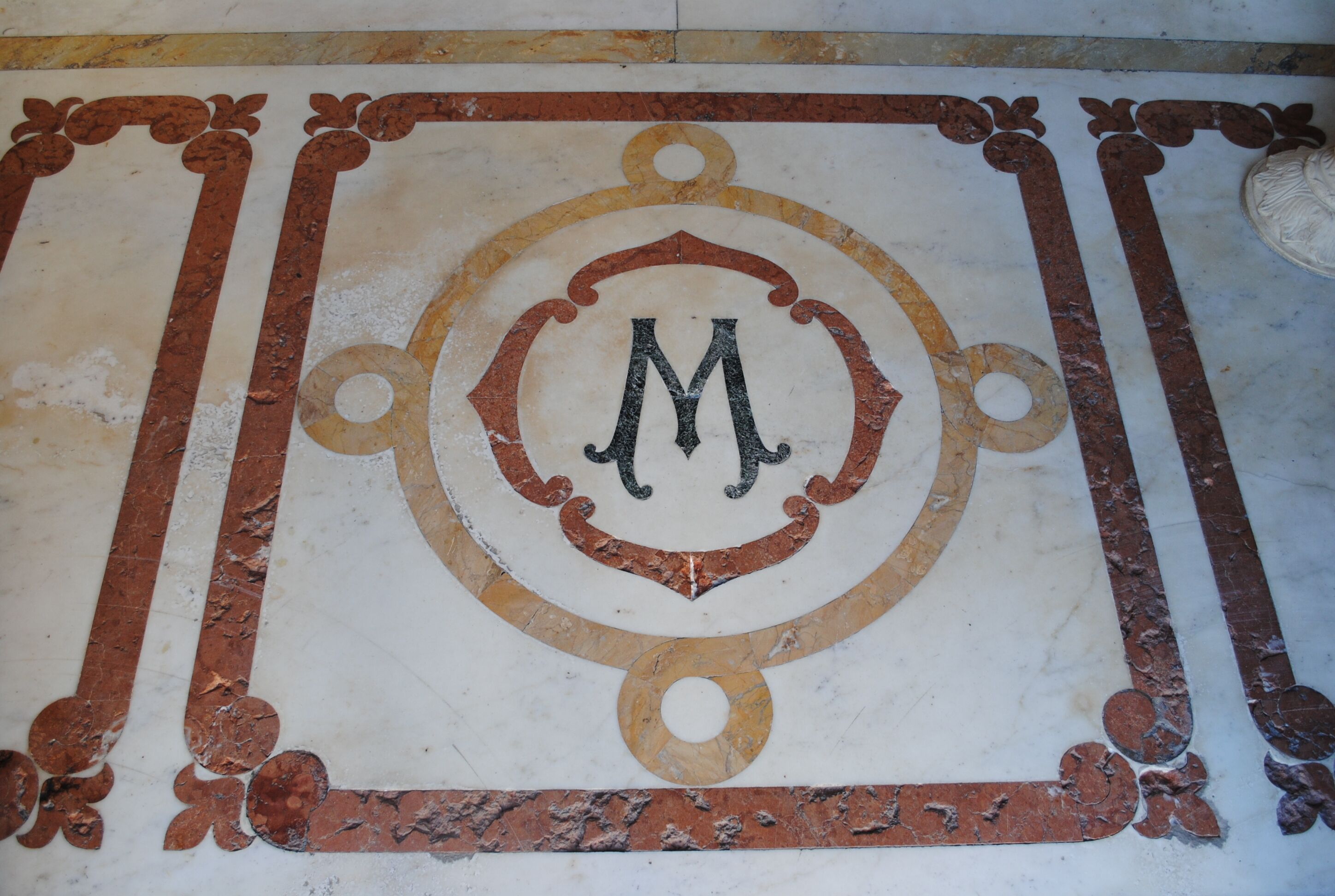 Un dettagliato mosaico pavimentale con uno stemma centrale raffigurante la lettera 'M' maiuscola.