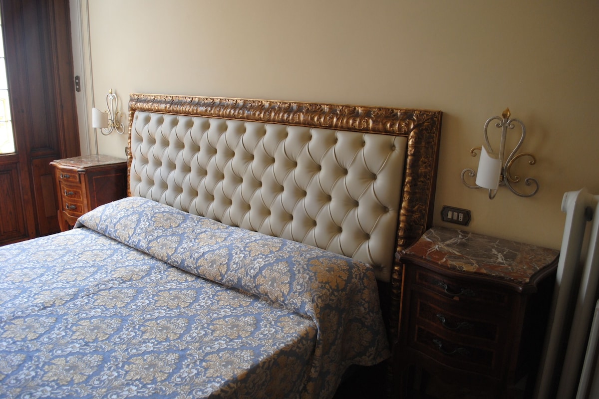 A bedroom with a tufted headboard, a patterned bedspread, a wooden nightstand, and a marble-topped dresser.