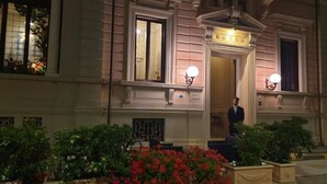 Entrada de un hotel con una fachada imponente, un hombre de pie en la puerta y un jardín bien cuidado con flores rojas.