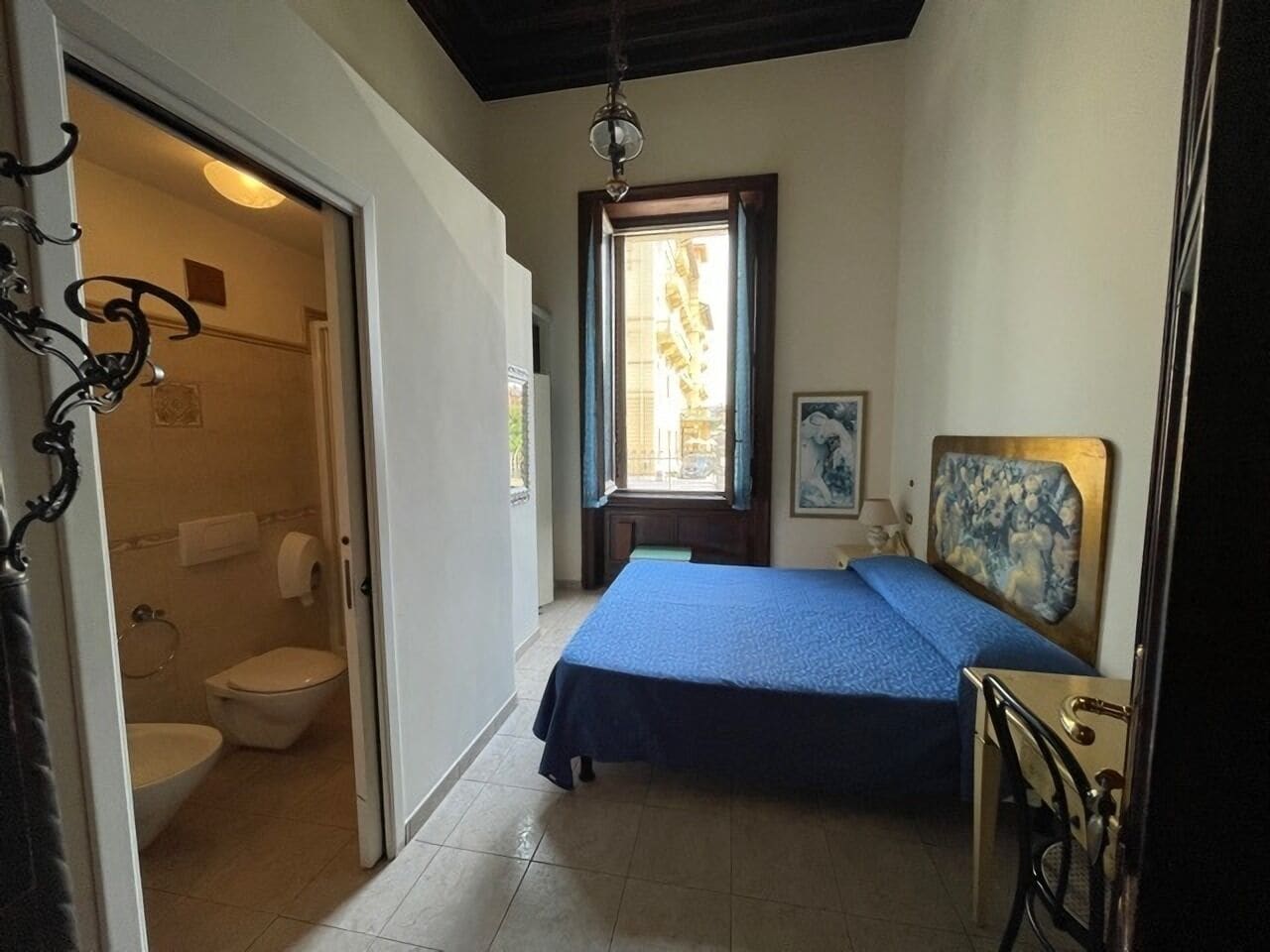 A bedroom with a bed, a window, a painting, and a bathroom visible through an open door.