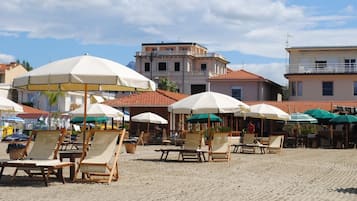 Relax on comfortable loungers under shaded umbrellas, surrounded by Mediterranean-style buildings and a sandy beach.