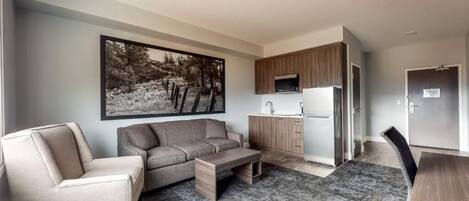 A modern living room with a sofa, armchair, and coffee table, a kitchen area with wooden cabinets and a microwave, and a large framed picture on the wall.