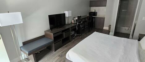 Una habitación de hotel con cama, televisor, escritorio con silla, banca y kitchenette.