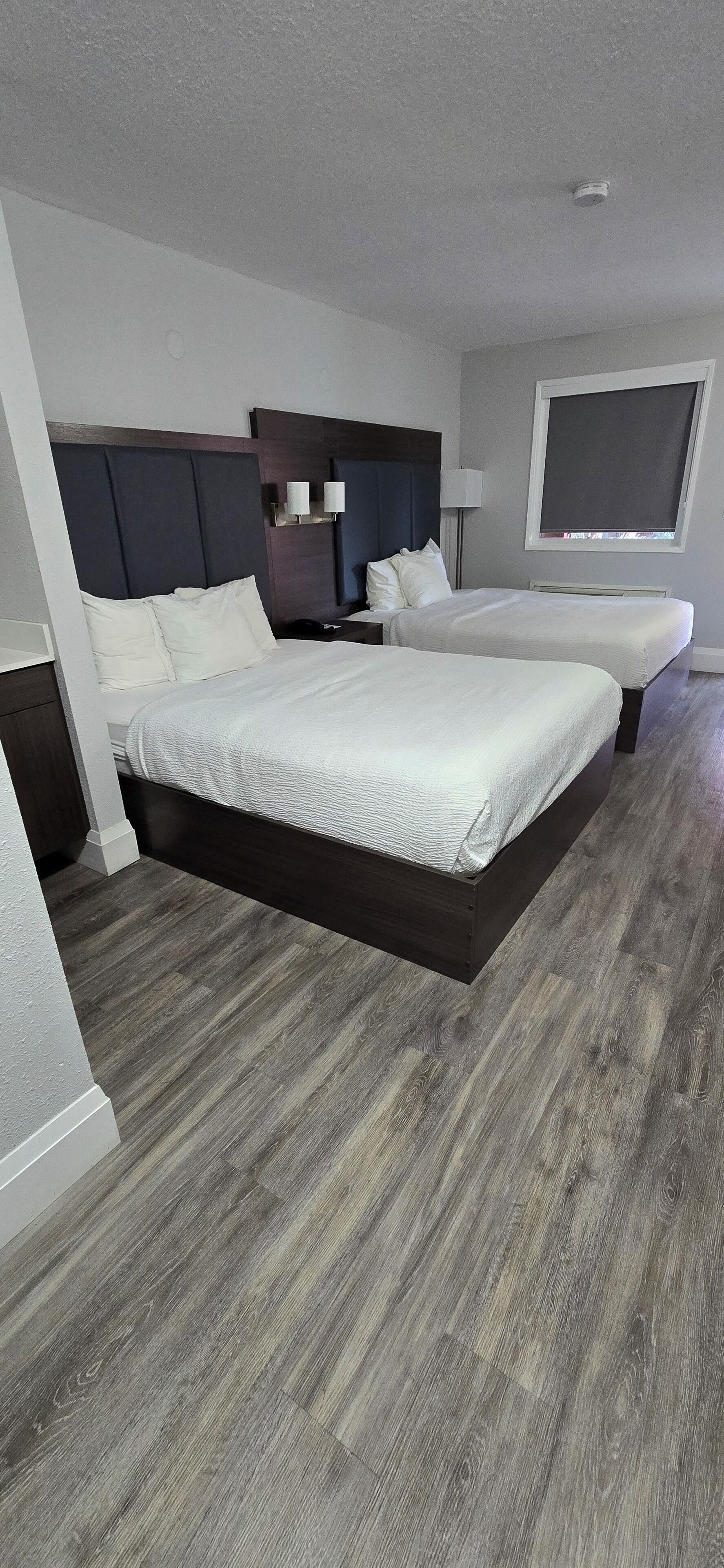 A modern hotel room with two beds, a dark wood floor, a large window, and a ceiling light.