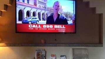 A television mounted on a wall displaying a news broadcast with a "CALL 898 BELL" advertisement. Below the TV, there is a wooden table with various items on it.