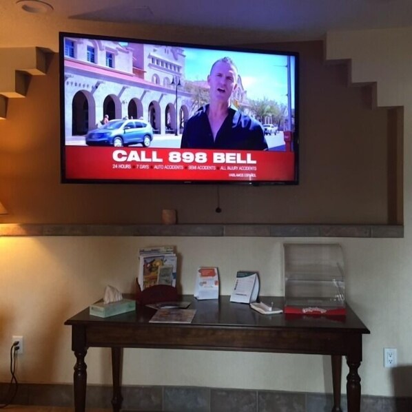 A television mounted on a wall displaying a news broadcast with a "CALL 898 BELL" advertisement. Below the TV, there is a wooden table with various items on it.