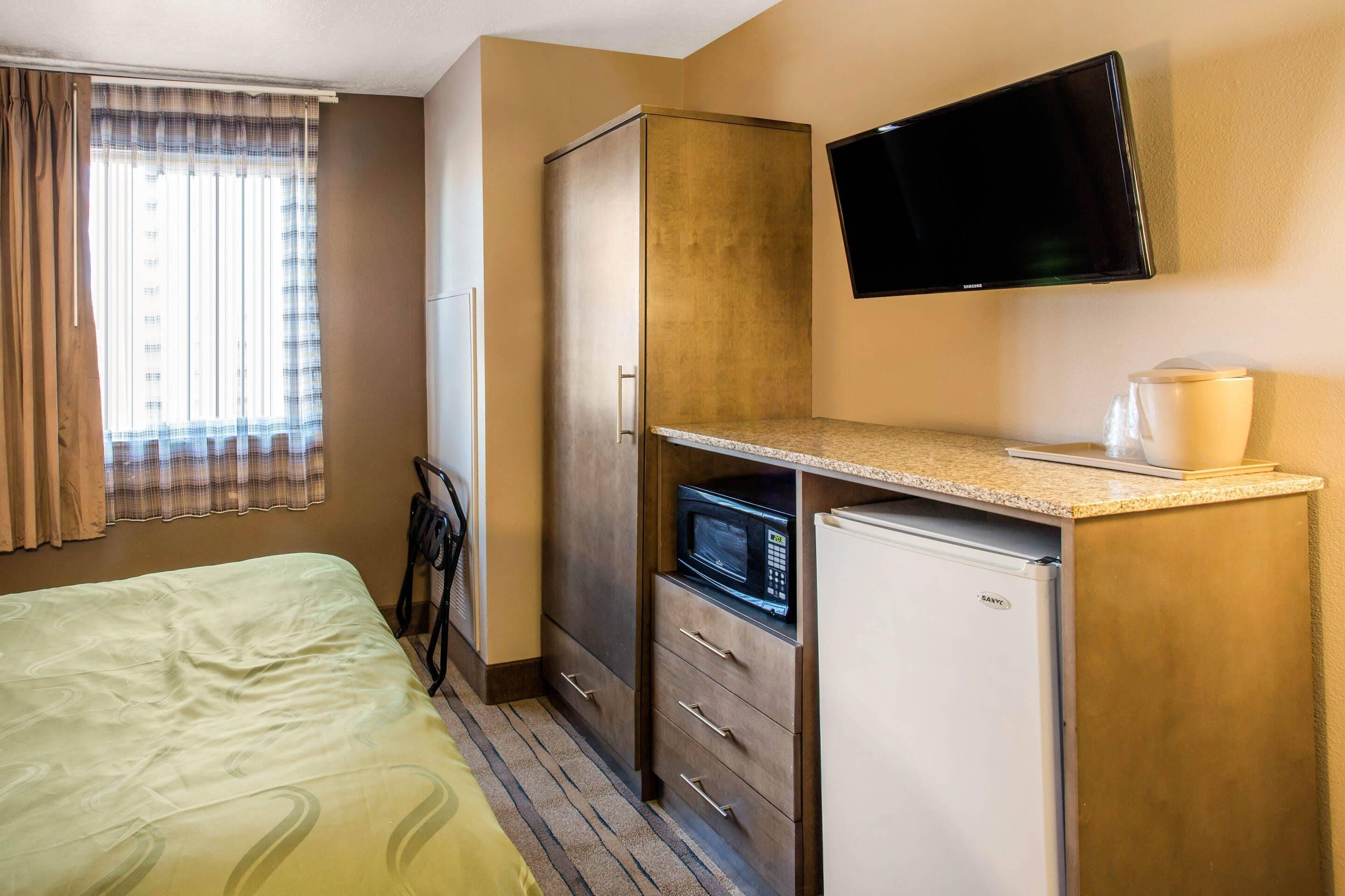 A hotel room with a bed, a kitchenette with a microwave, a refrigerator, a flat-screen TV mounted on the wall, and a window with curtains.