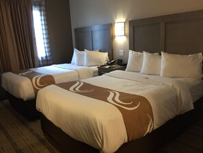 A hotel room with two beds, each with white bedding and a brown accent design. There is a nightstand with a phone and a lamp between the beds.